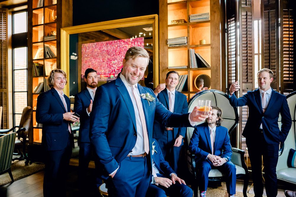Groomsmen in navy suits posing against pink floral wall in modern venue.