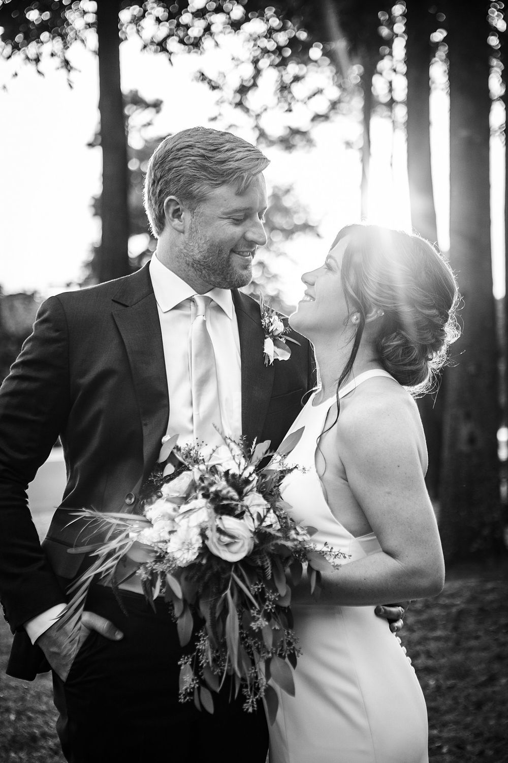 Black and white portrait of newlyweds sharing intimate moment in sunlit forest.