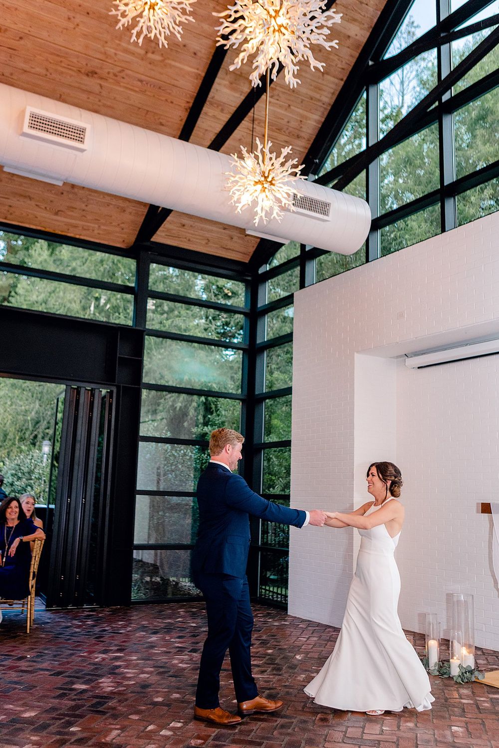 Couple dances under stunning starburst chandeliers in modern venue with floor-to-ceiling windows.