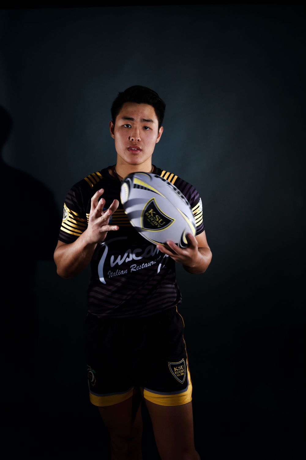 Rugby player in black and yellow striped uniform poses with ball against dark background.
