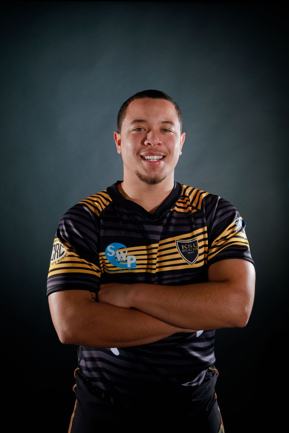 Rugby player in black and gold striped jersey poses confidently against dark background.