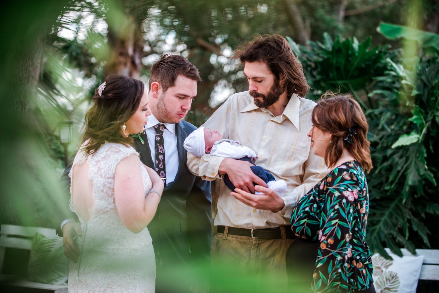 Wedding guests gather around a newborn baby in a lush garden setting.