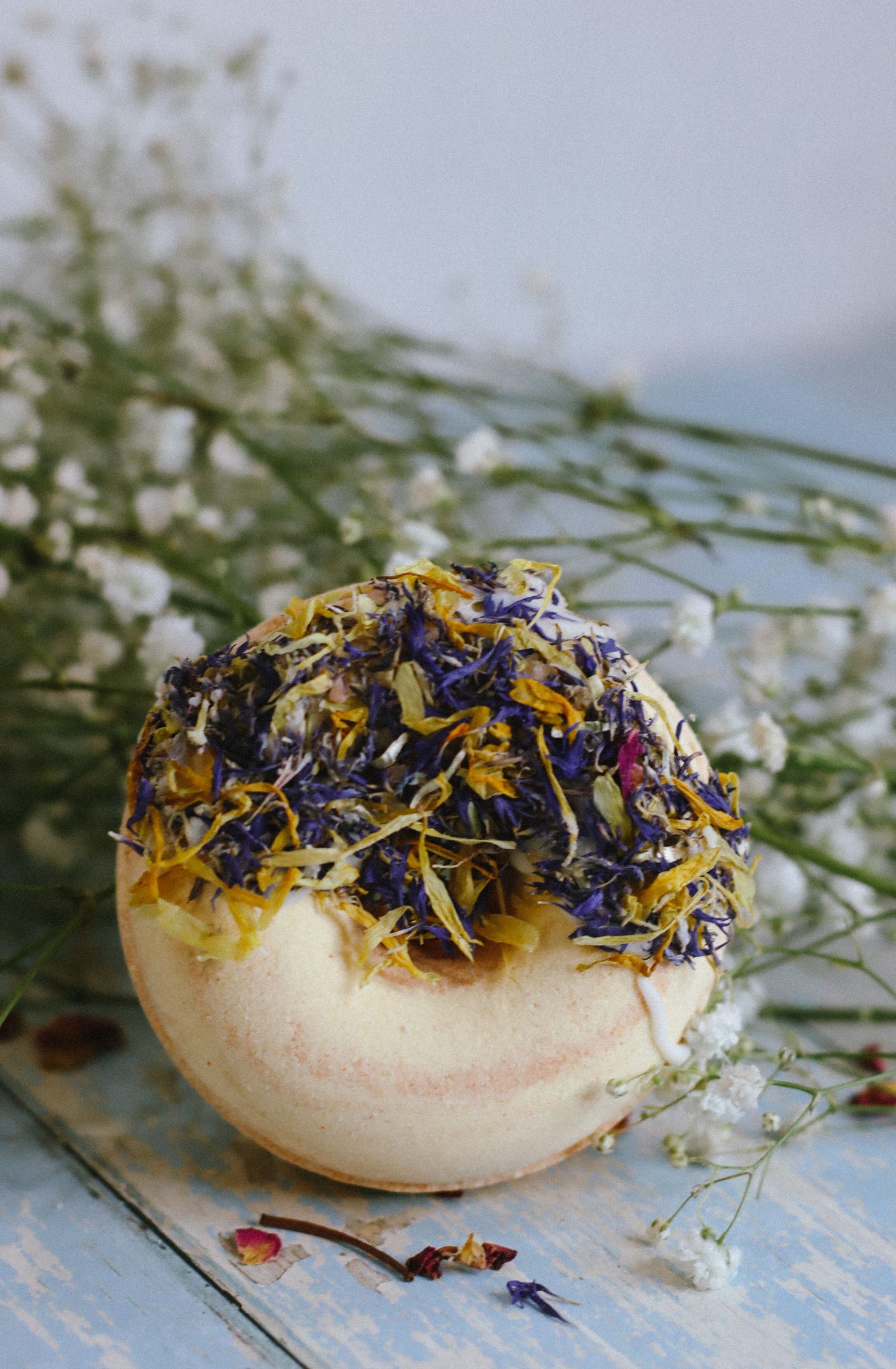Bath bomb decorated with dried yellow and purple flower petals on a rustic blue surface.