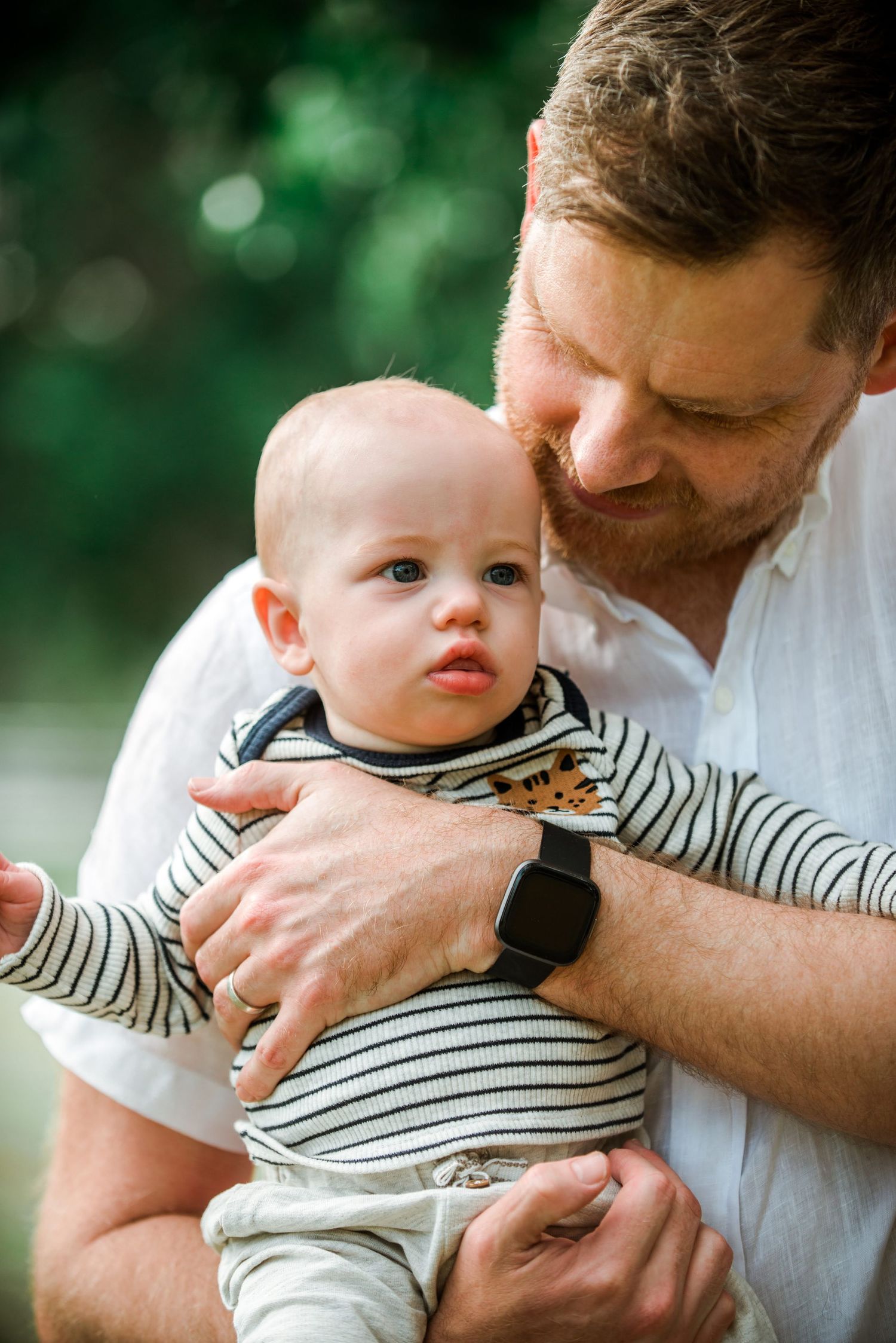Father in white shirt embracing baby in striped outfit with green bokeh background.
