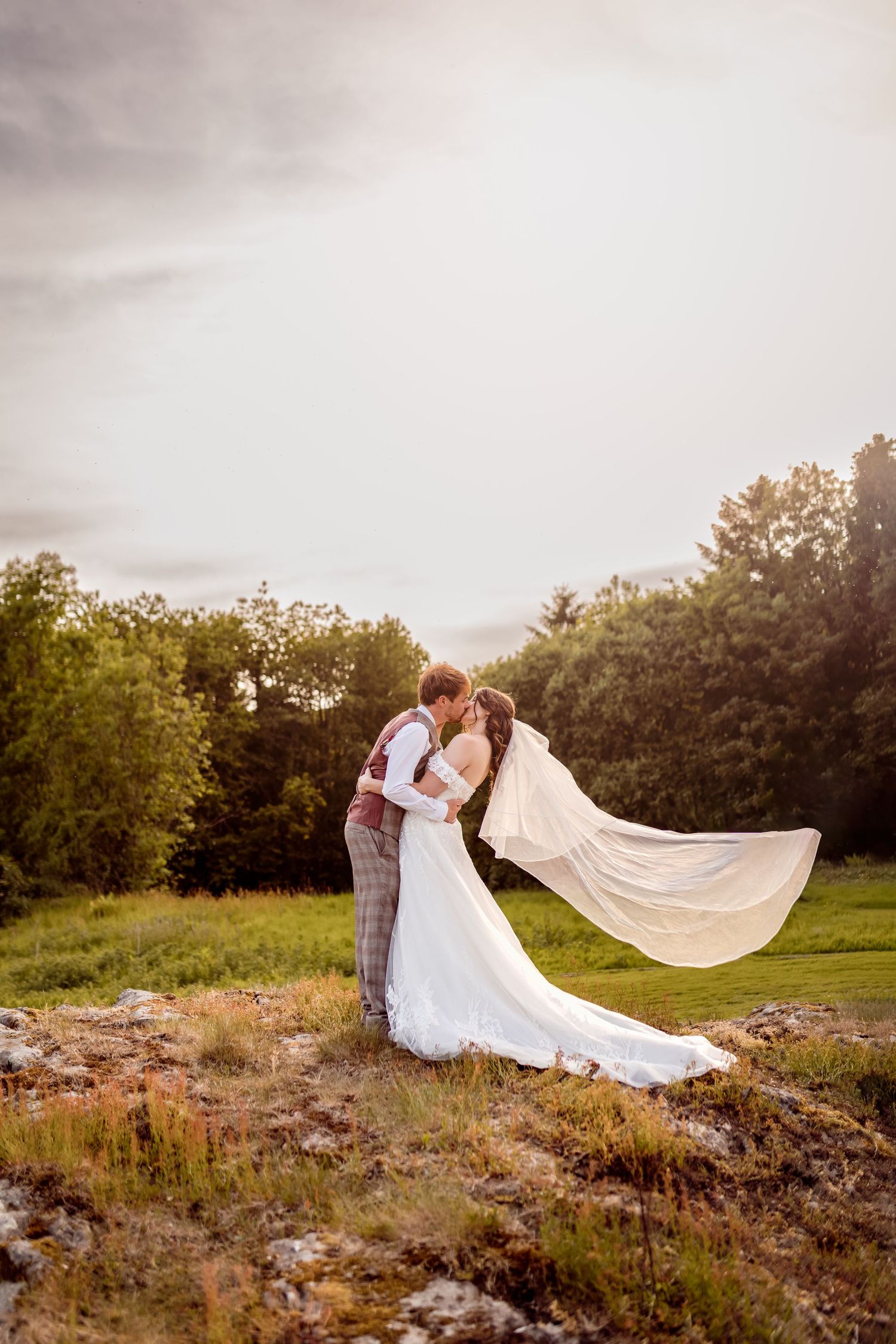 Couple in wedding attire share an intimate moment as bride's veil flows in the wind.