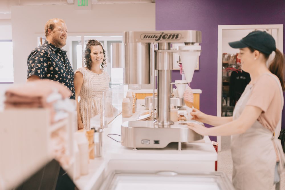 A woman stands at a little Jem ice cream machine holding a cone infront of counter a couple looks at woman