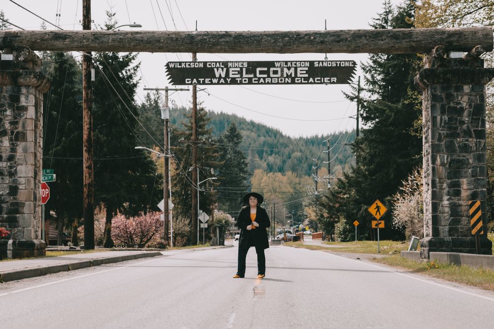 A woman stands in the middle of the street underneath a wooden sign that says Welcome