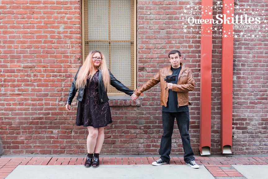 A couple poses against a vibrant red brick wall in an outdoor engagement photo session.