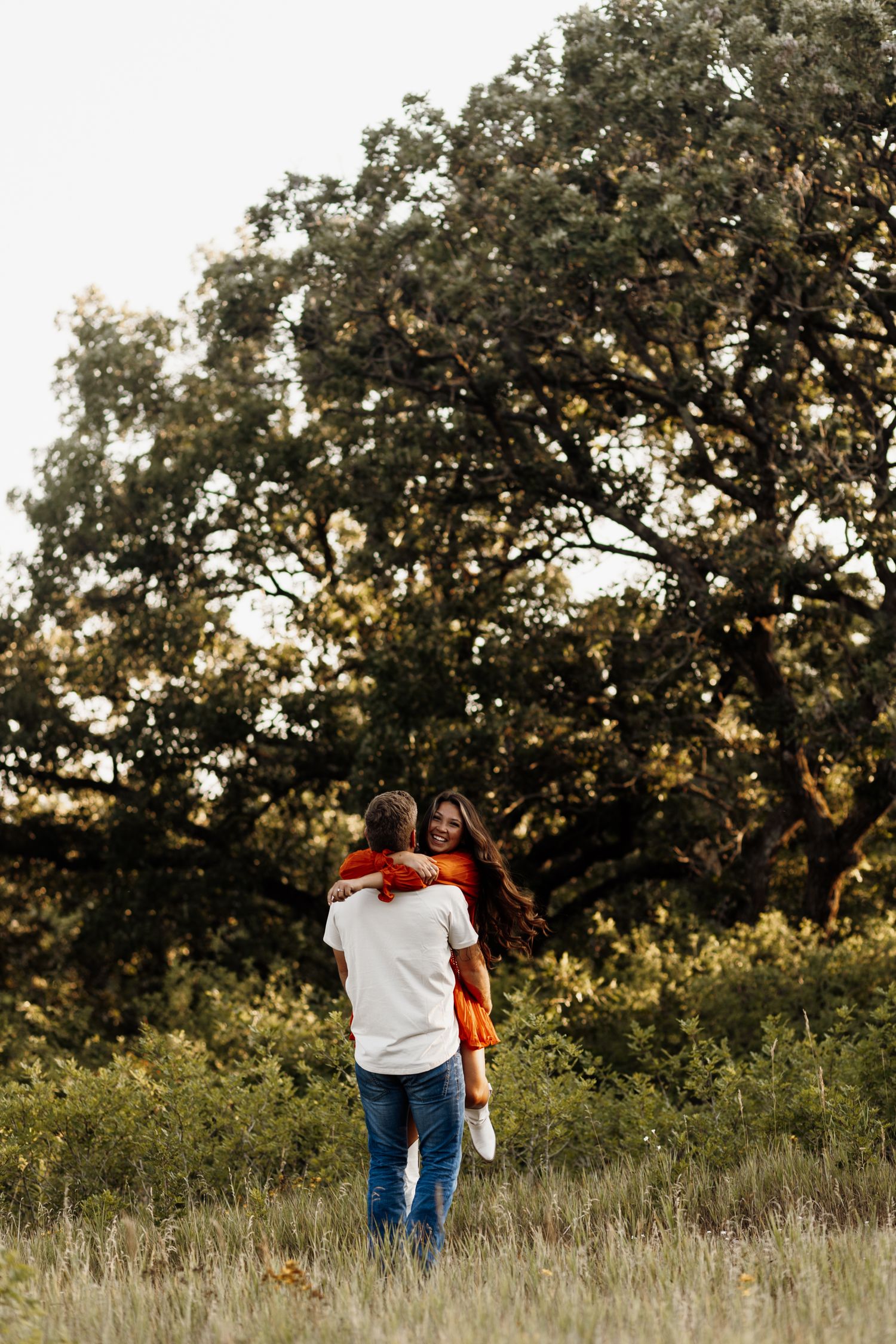 Couple walking through tall grass towards a majestic oak tree at dusk.