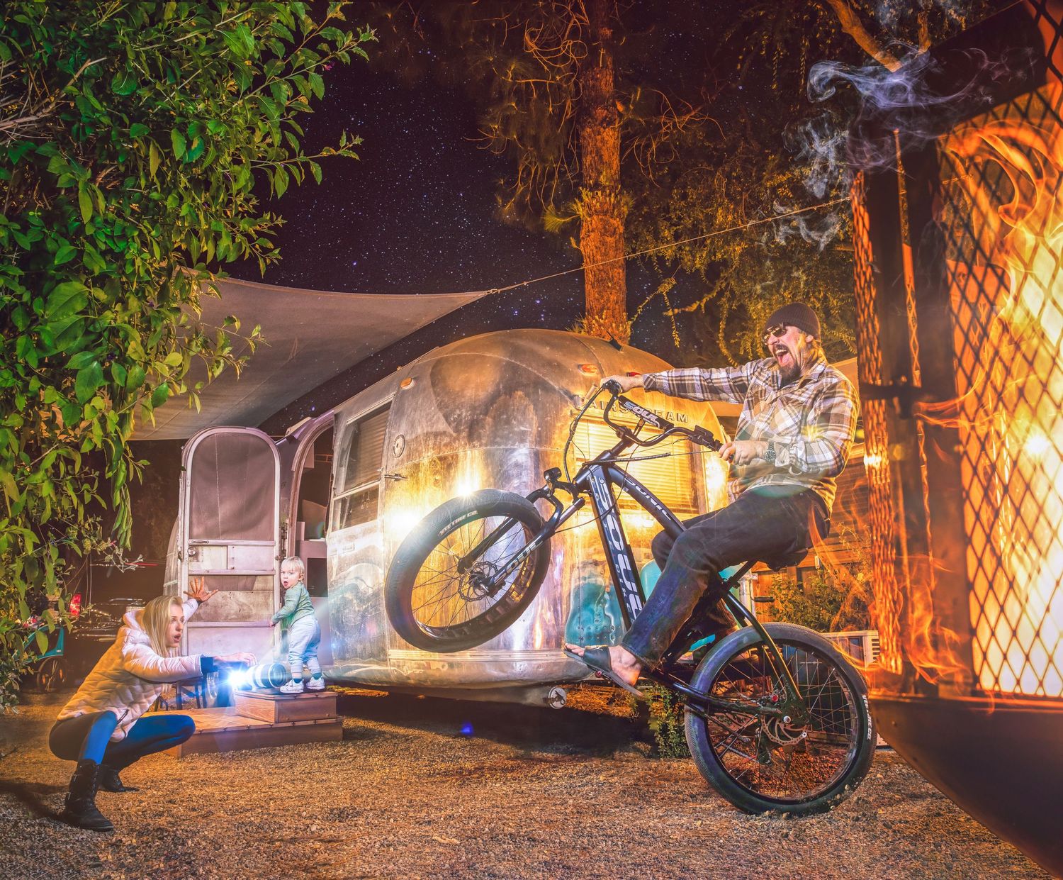 Man performing a wheelie on an electric bike at night near an Airstream trailer with light trails.