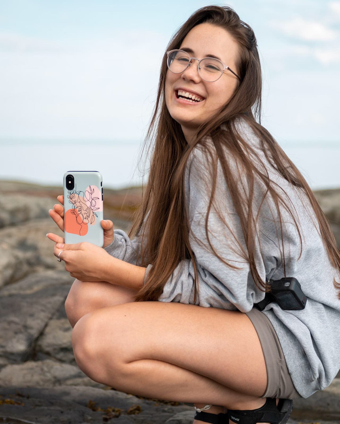 Smiling young woman with glasses holding a floral phone case while crouching on rocks near the ocean.