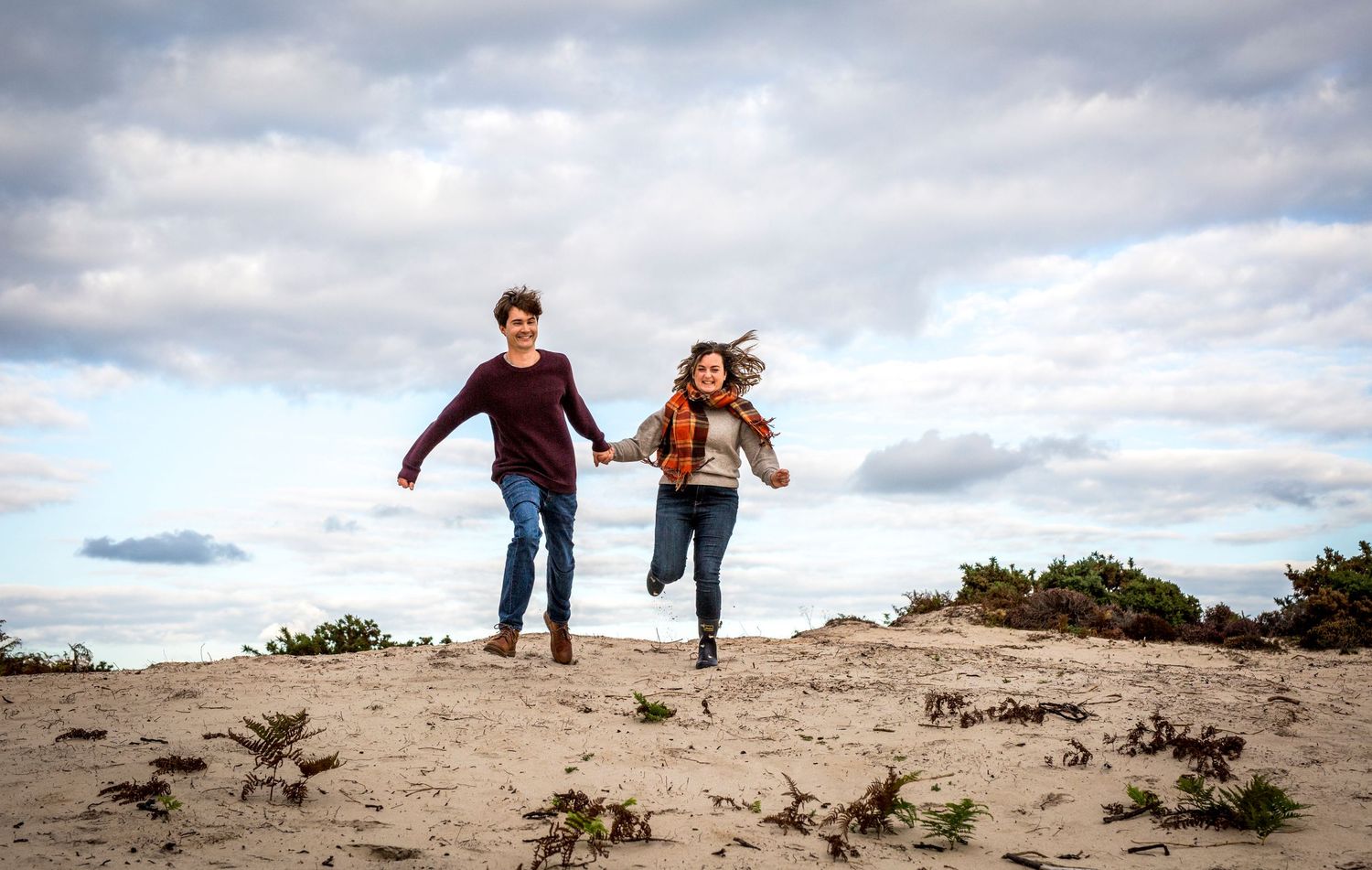 Two people running and playing on sandy dunes under dramatic cloudy skies.