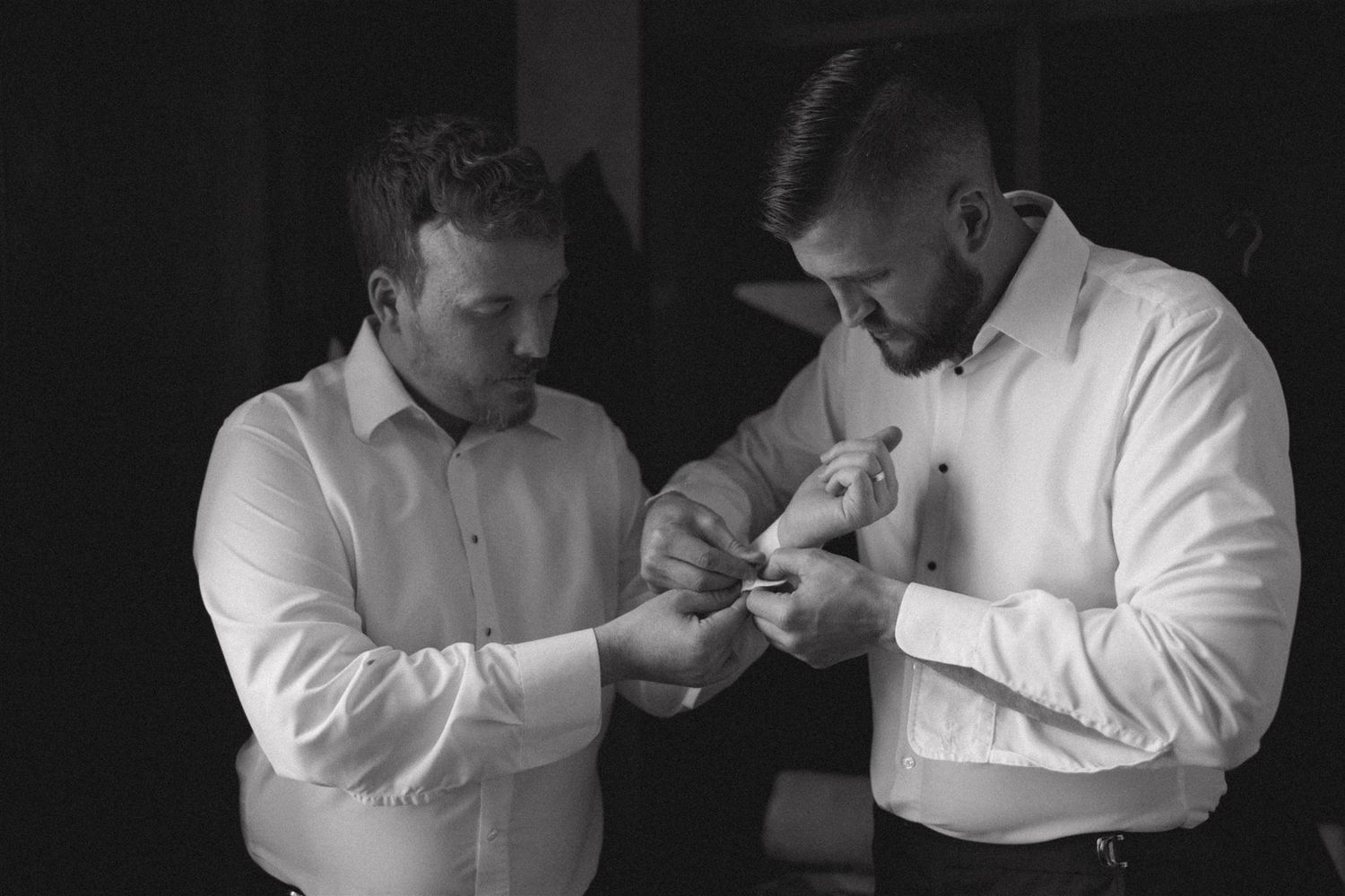 Two people in white dress shirts help each other with cuff links or buttons in a black and white photograph.