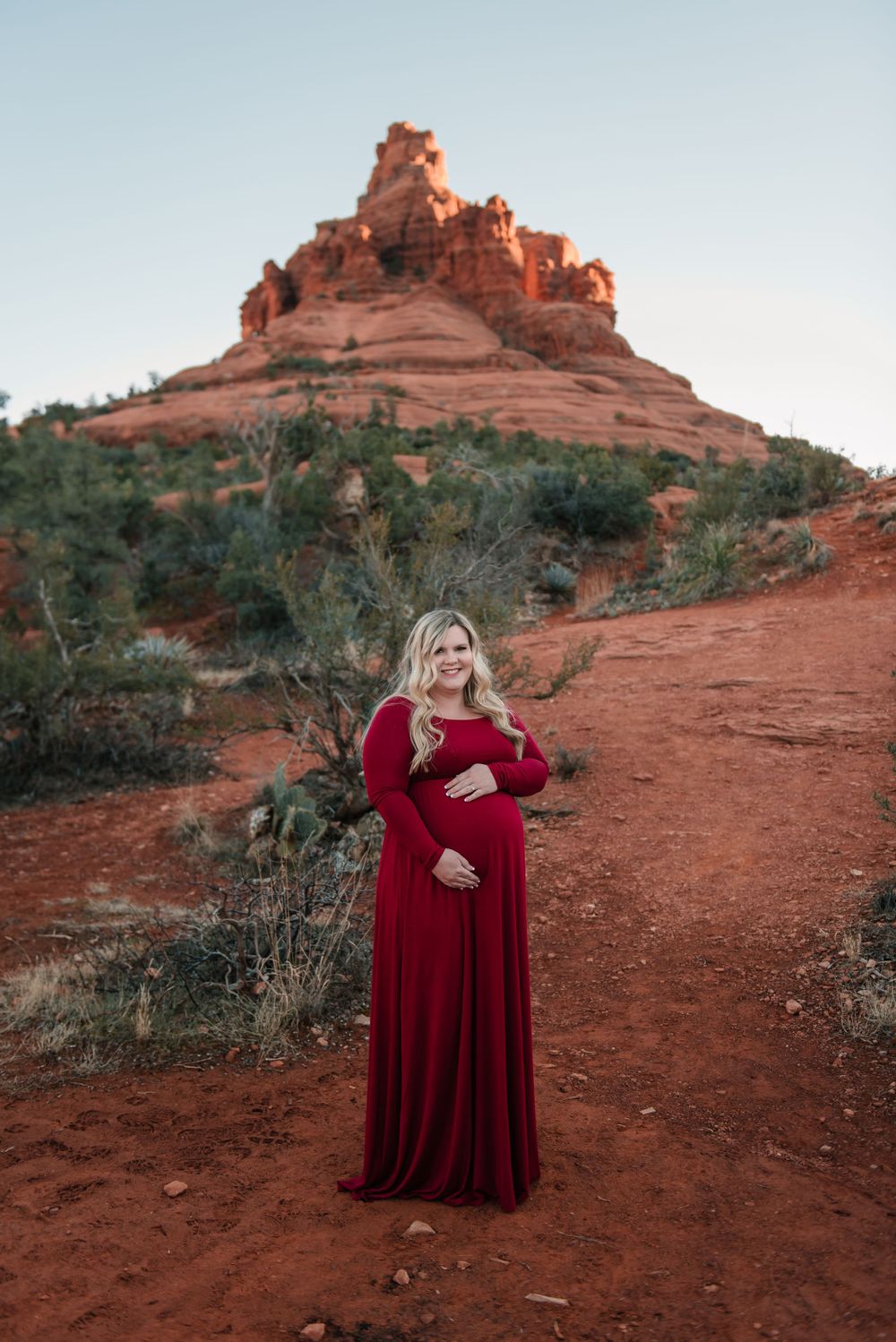 A person in a burgundy dress stands on red rock terrain with desert vegetation and mountains behind.