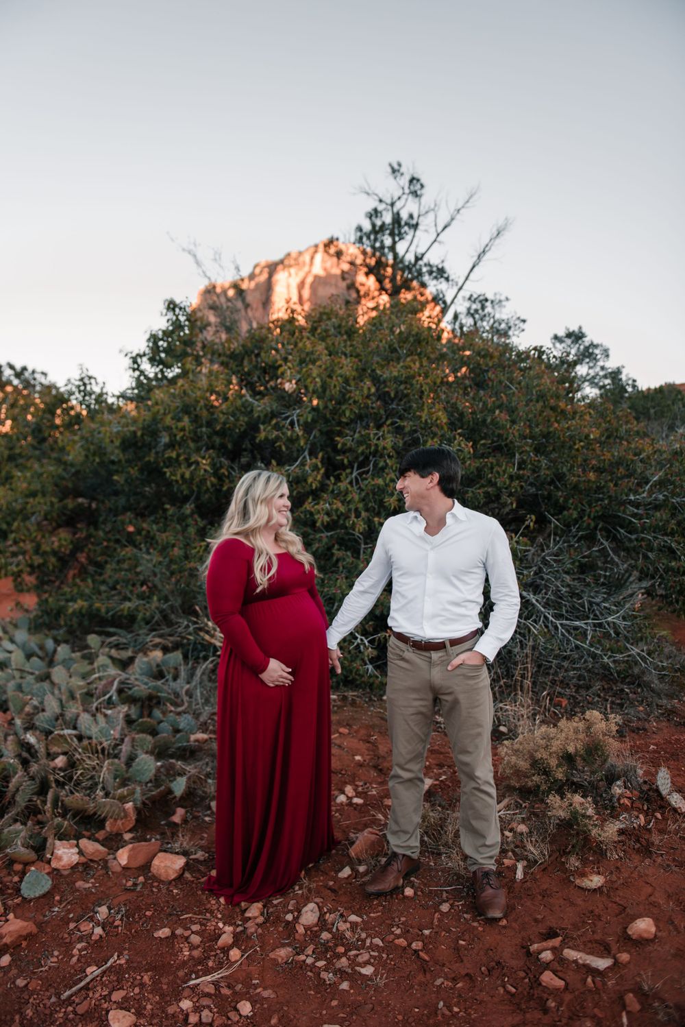 A couple shares a romantic moment against a backdrop of red rocks and desert brush in an outdoor photoshoot.