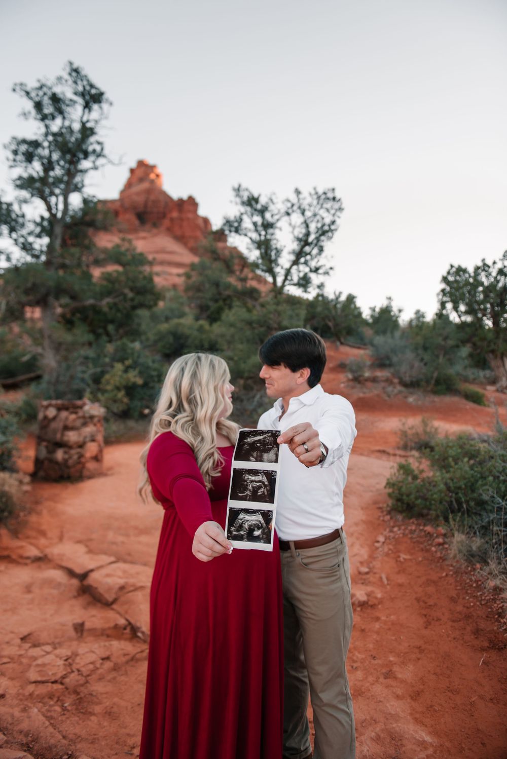 A couple holds up an ultrasound photo while standing on red desert terrain with rock formations in the background.