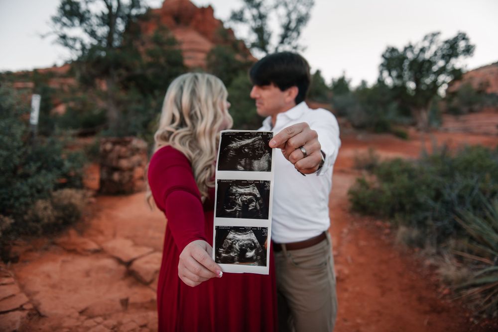 A couple embraces while displaying a sonogram photo in a desert landscape with red rocks.