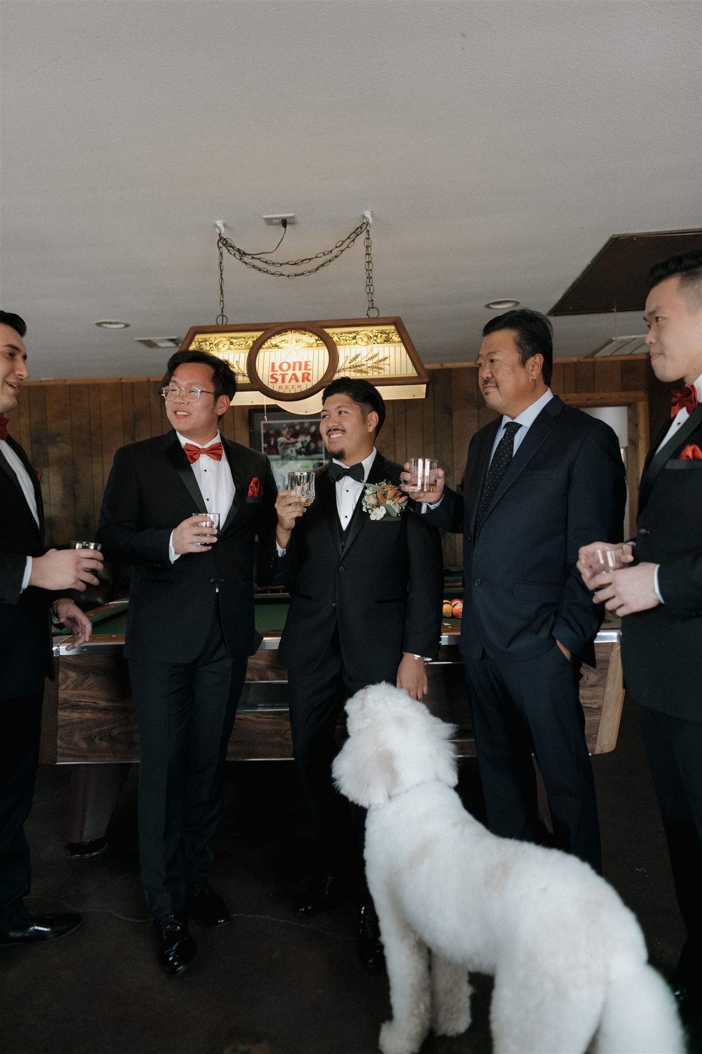 Groomsmen socializing with drinks while a white dog joins their pre-wedding celebration.
