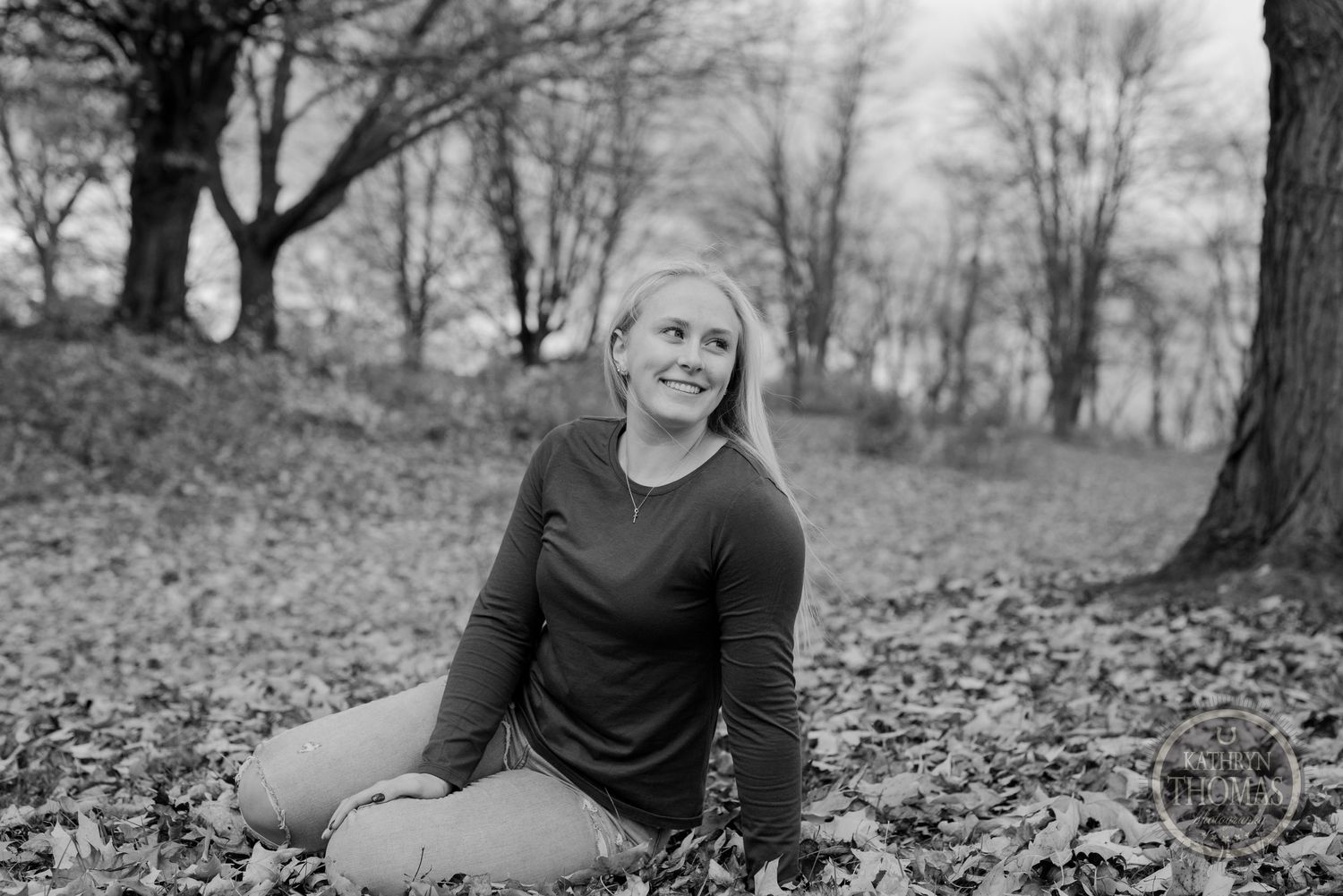 Person sitting on autumn leaves wearing dark sweater in black and white forest setting.