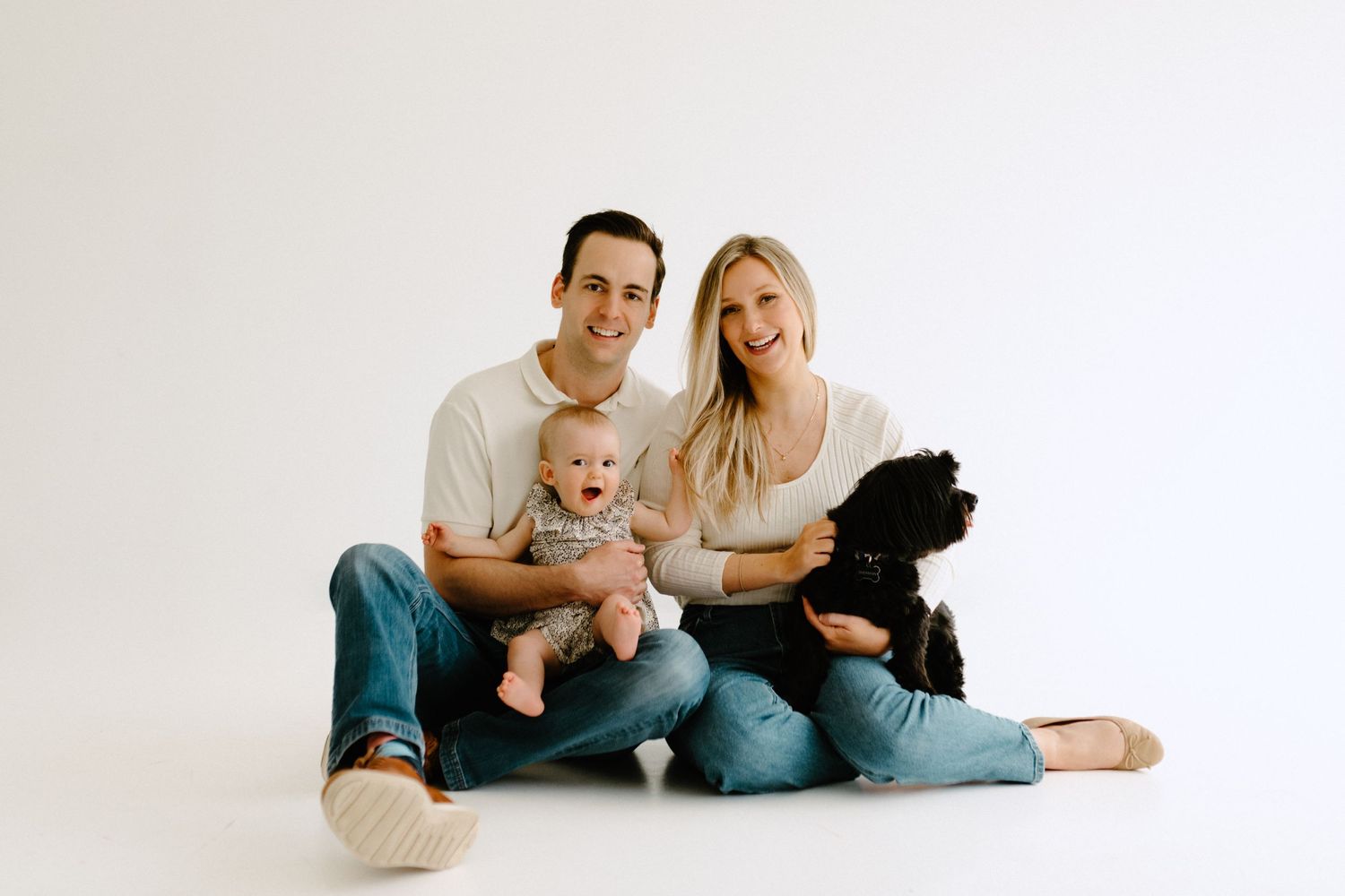 A minimalist studio portrait of a family with their black dog against a white background.