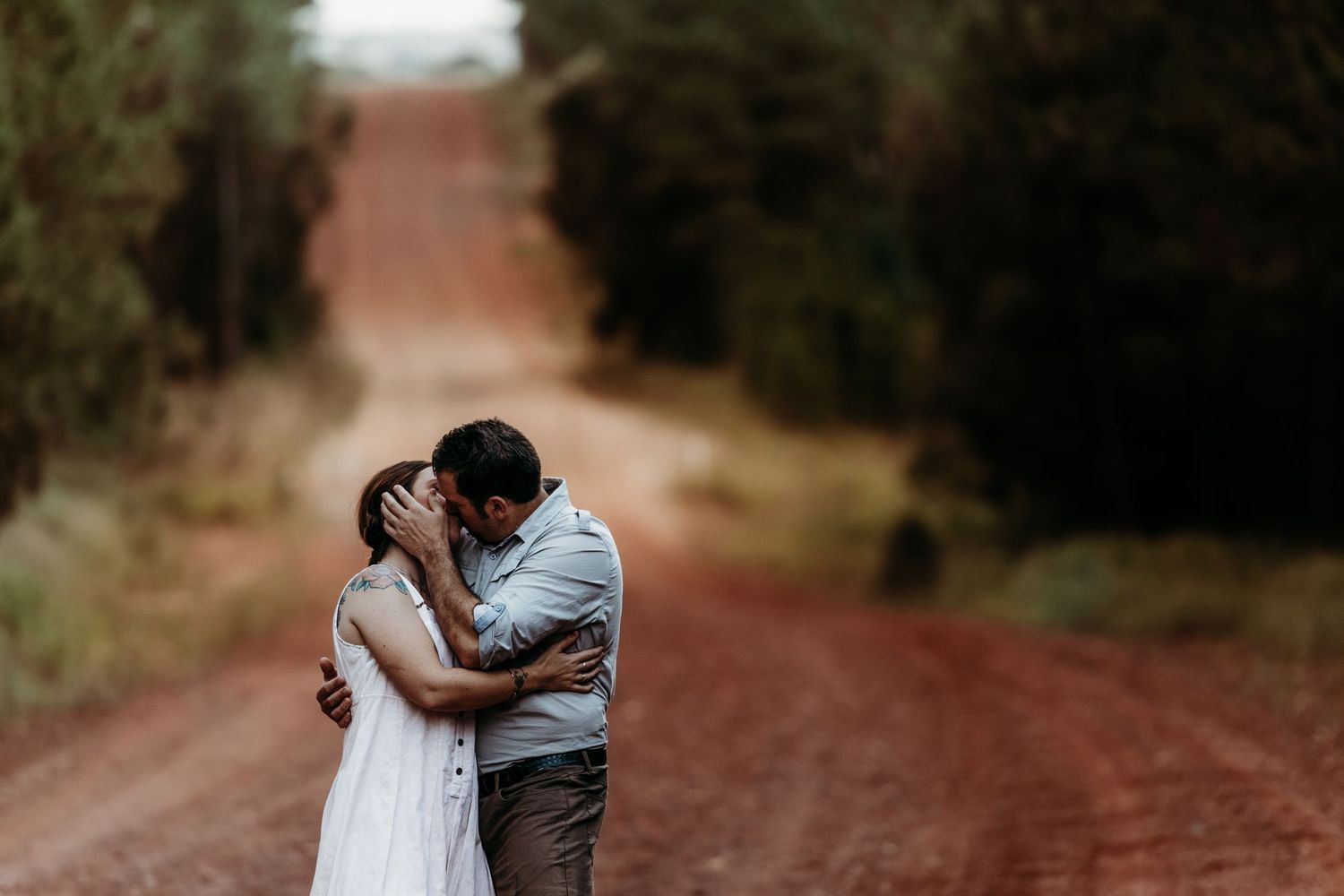 A couple shares a kiss on a red dirt road surrounded by tall pine trees.