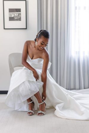 Wedding dress detail showing elegant white gown and strappy heels on a white floor.