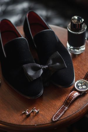 Groom's accessories including black velvet loafers with bow tie, watch, and cufflinks on wooden surface.