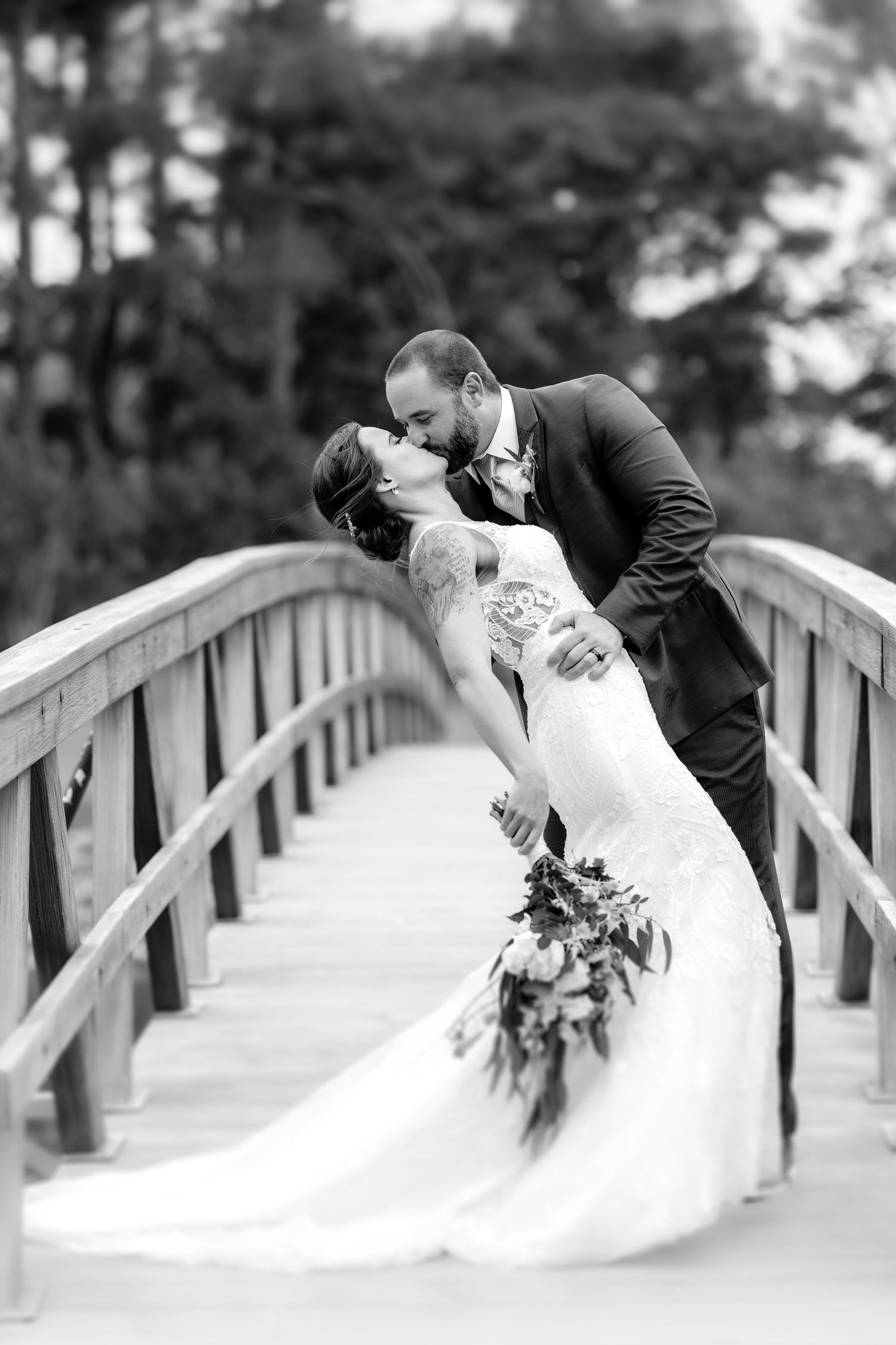 Romantic kiss between newlyweds on a wooden bridge with flowing wedding dress and bouquet.