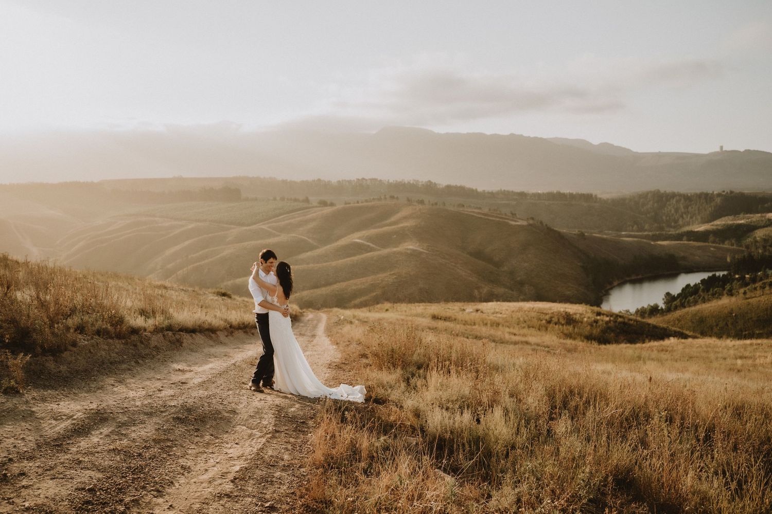 Wedding couple stands on hilltop path overlooking scenic valley at sunset.