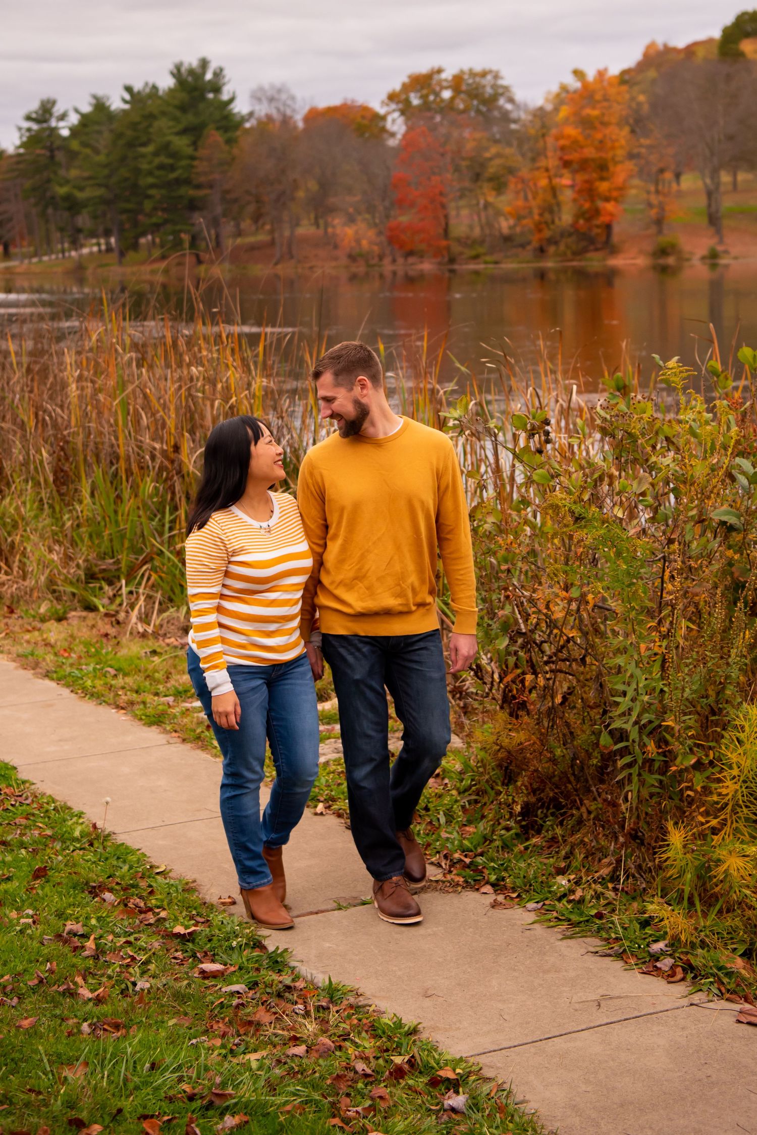 husband and wife walking along path holding hands with fall trees at north park lake