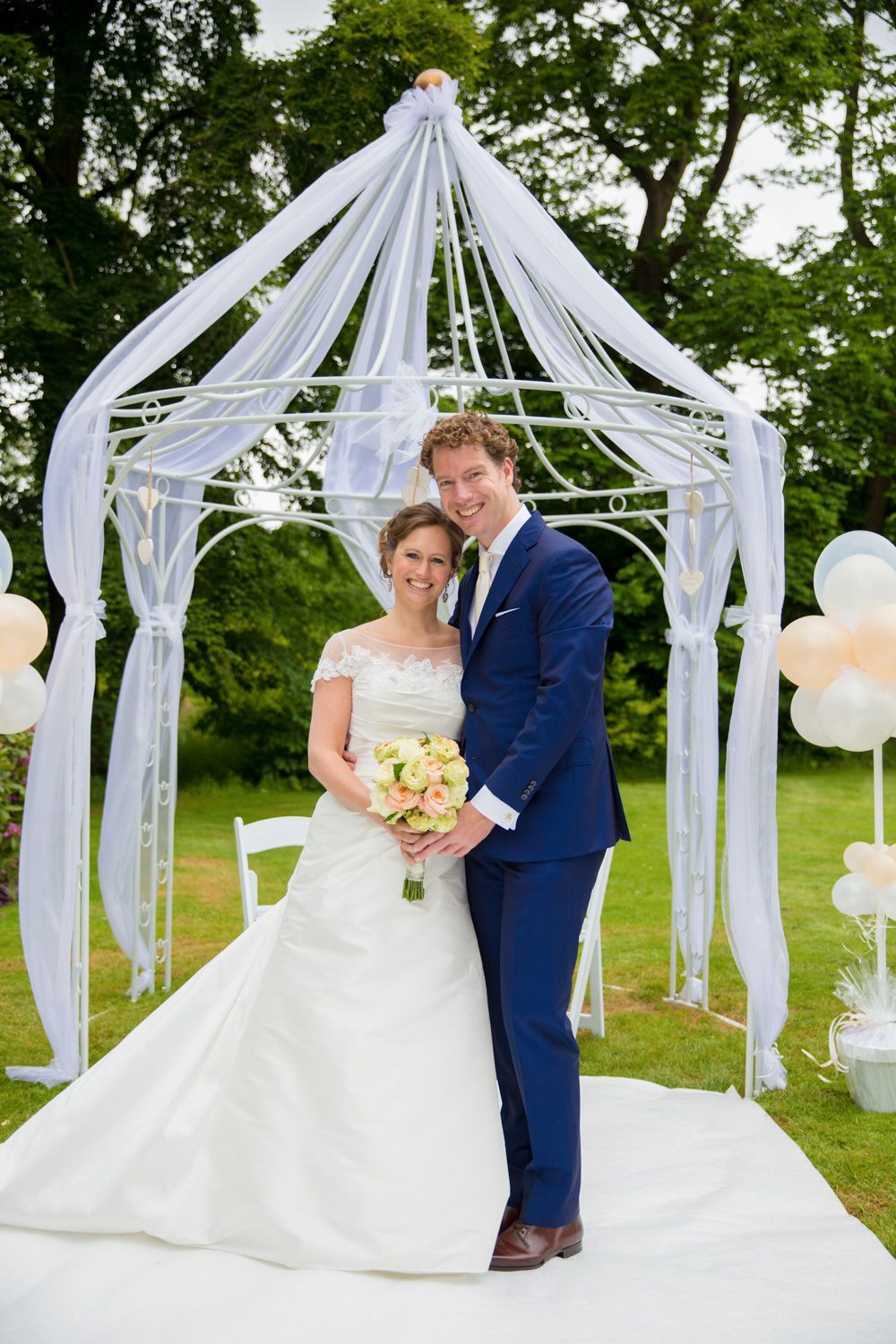 Newlyweds pose under white draped gazebo with floral bouquet.