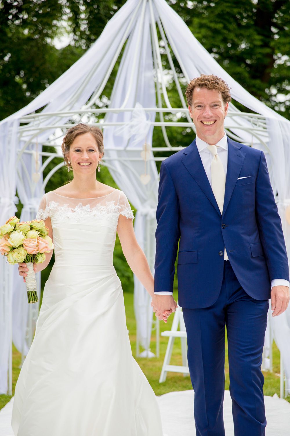 Wedding couple stands in formal attire under decorated white gazebo.