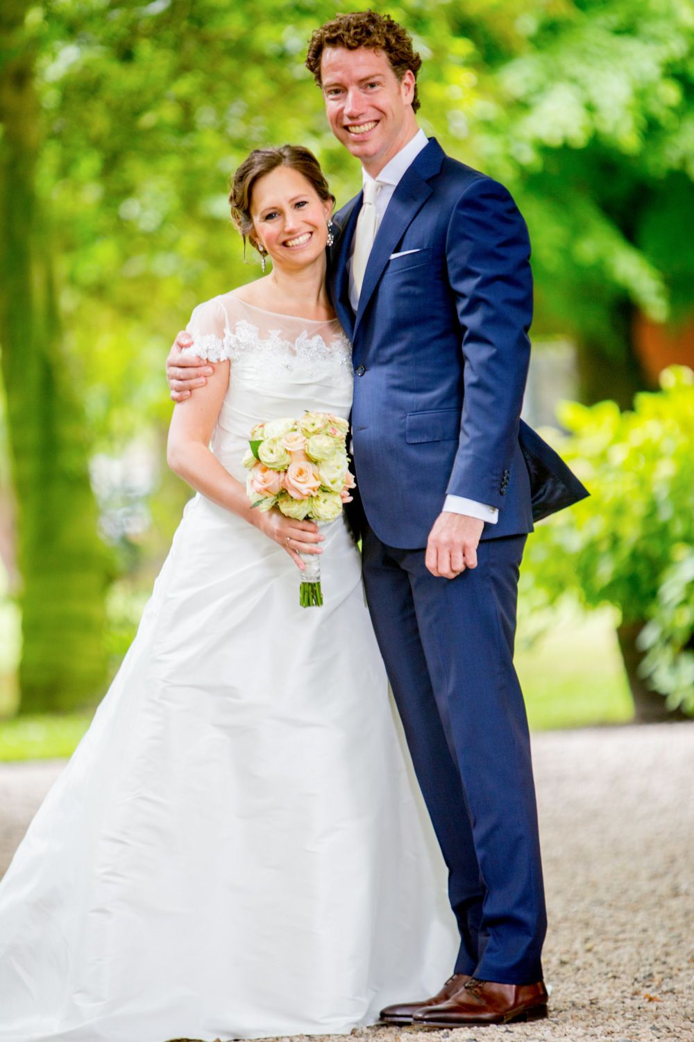 A bride in a white gown holds a bouquet of flowers while standing with her groom in a navy suit outdoors.