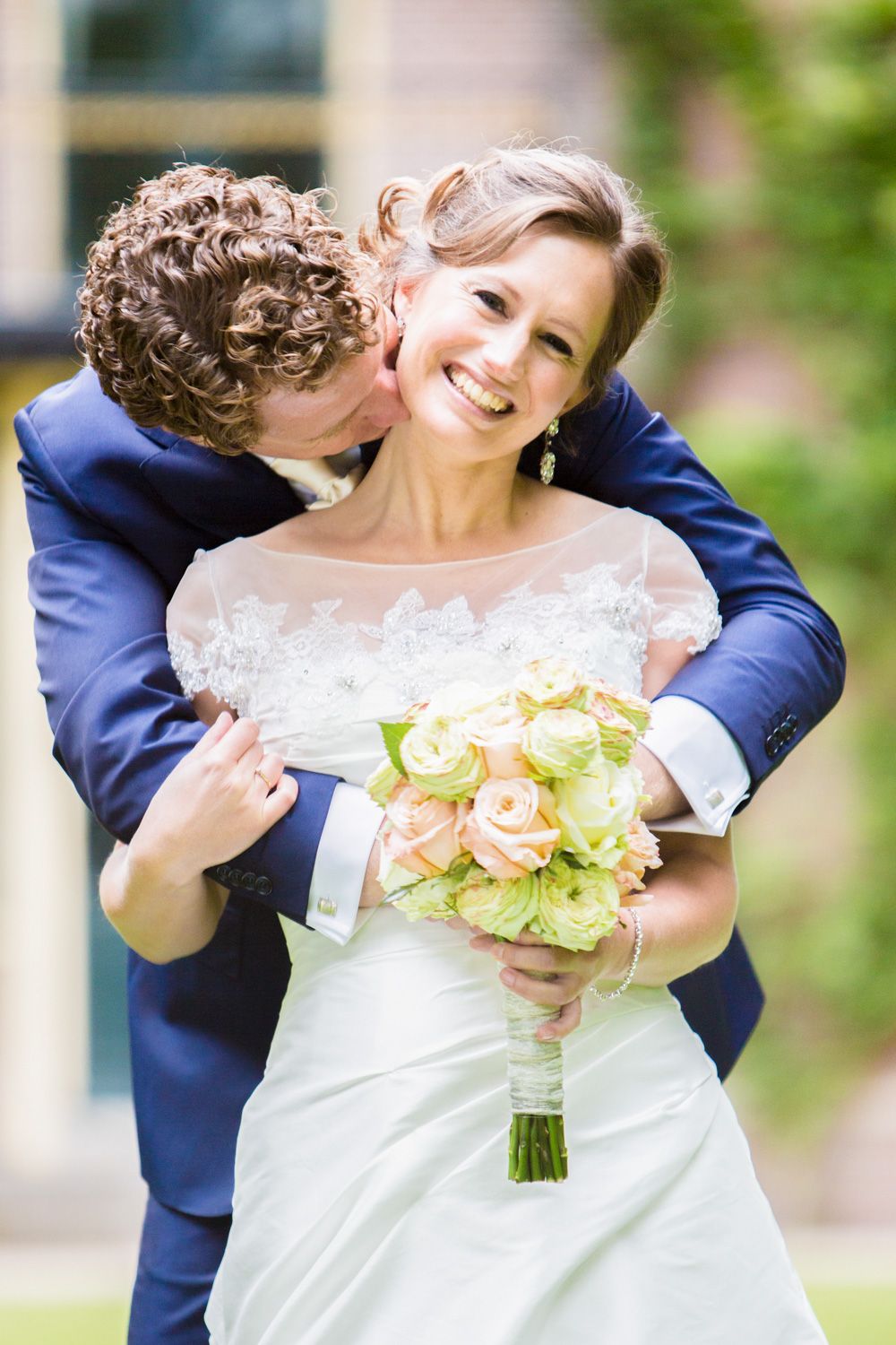 Intimate moment between newlyweds with white and peach bouquet against brick wall.