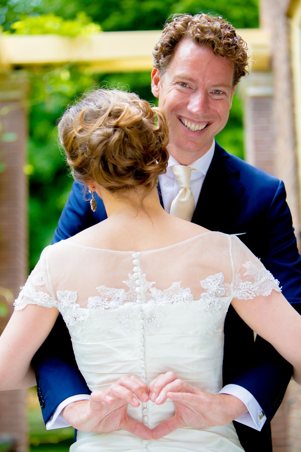 Wedding couple creates heart shape with hands behind bride's back in garden setting.