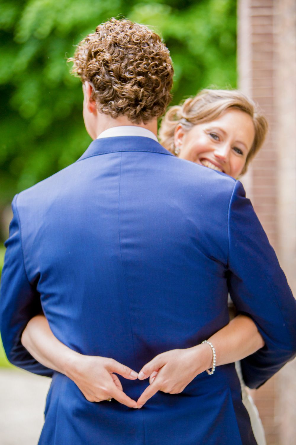 Wedding couple embraces while making heart shape with hands behind groom's back.