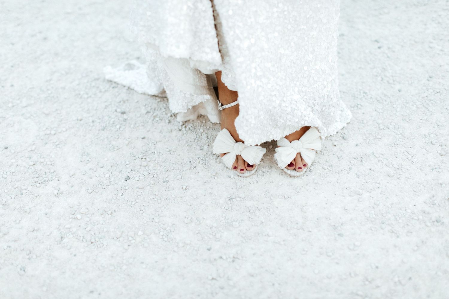 Close-up of bridal shoes peeking beneath white wedding dress on light colored ground.