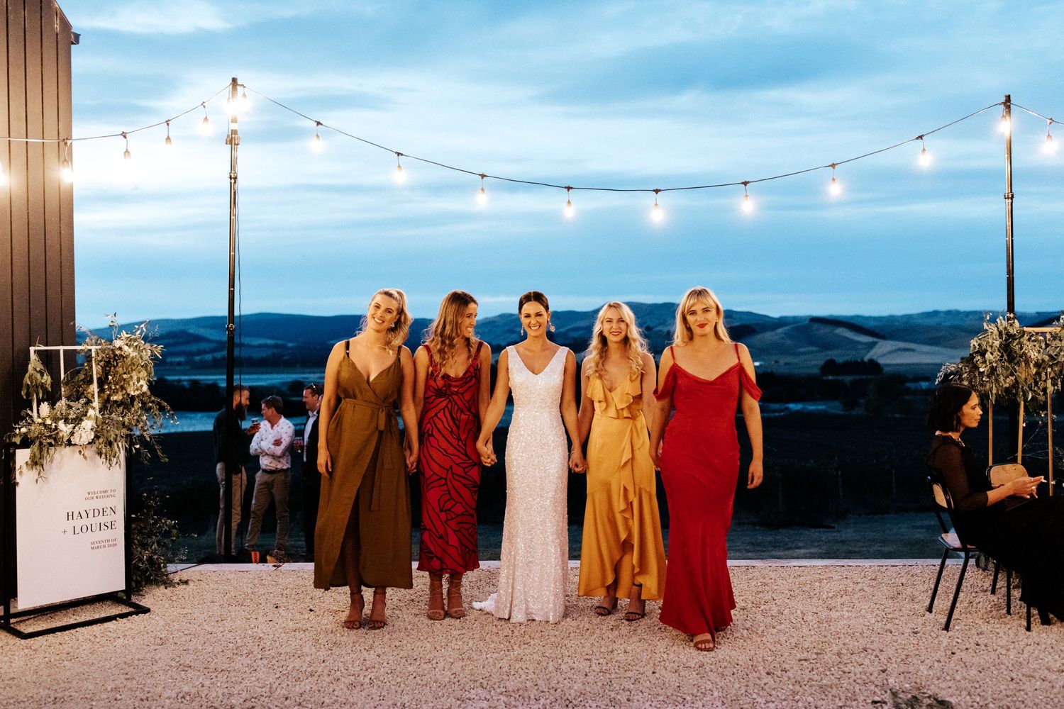 Wedding party poses against sunset sky with string lights and mountain views.