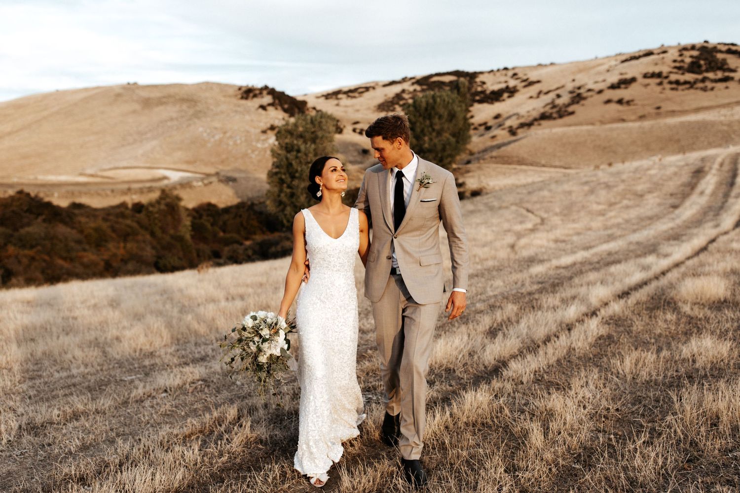 Wedding couple walks hand in hand through golden grass on scenic hillside.