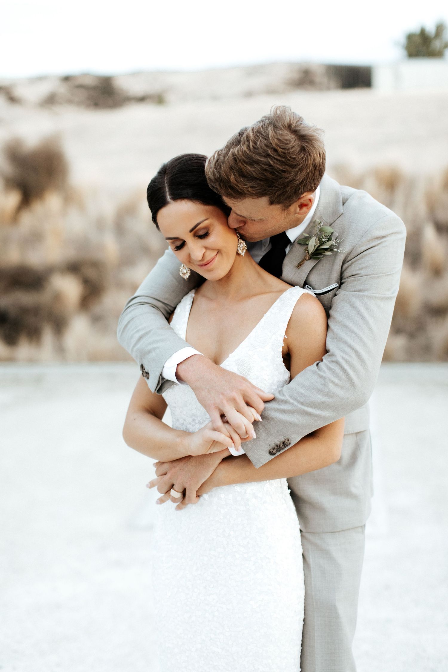 Romantic wedding couple embraces on a desert path wearing elegant white dress and light gray suit.