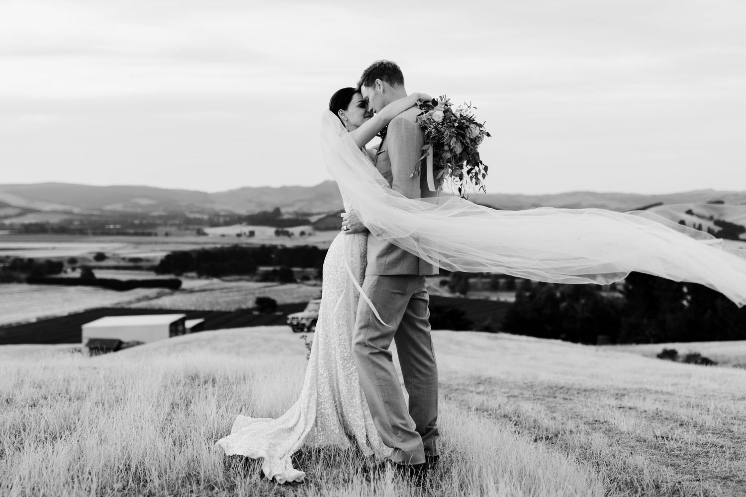 Black and white wedding photo capturing joyful embrace against rolling hills landscape.