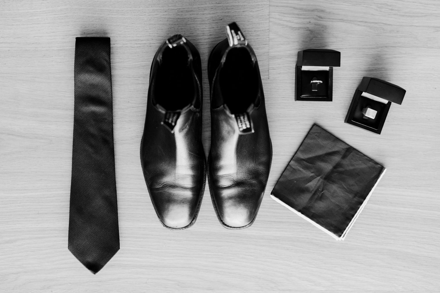 Wedding accessories flatlay with black dress shoes, tie, and ring boxes on light surface.