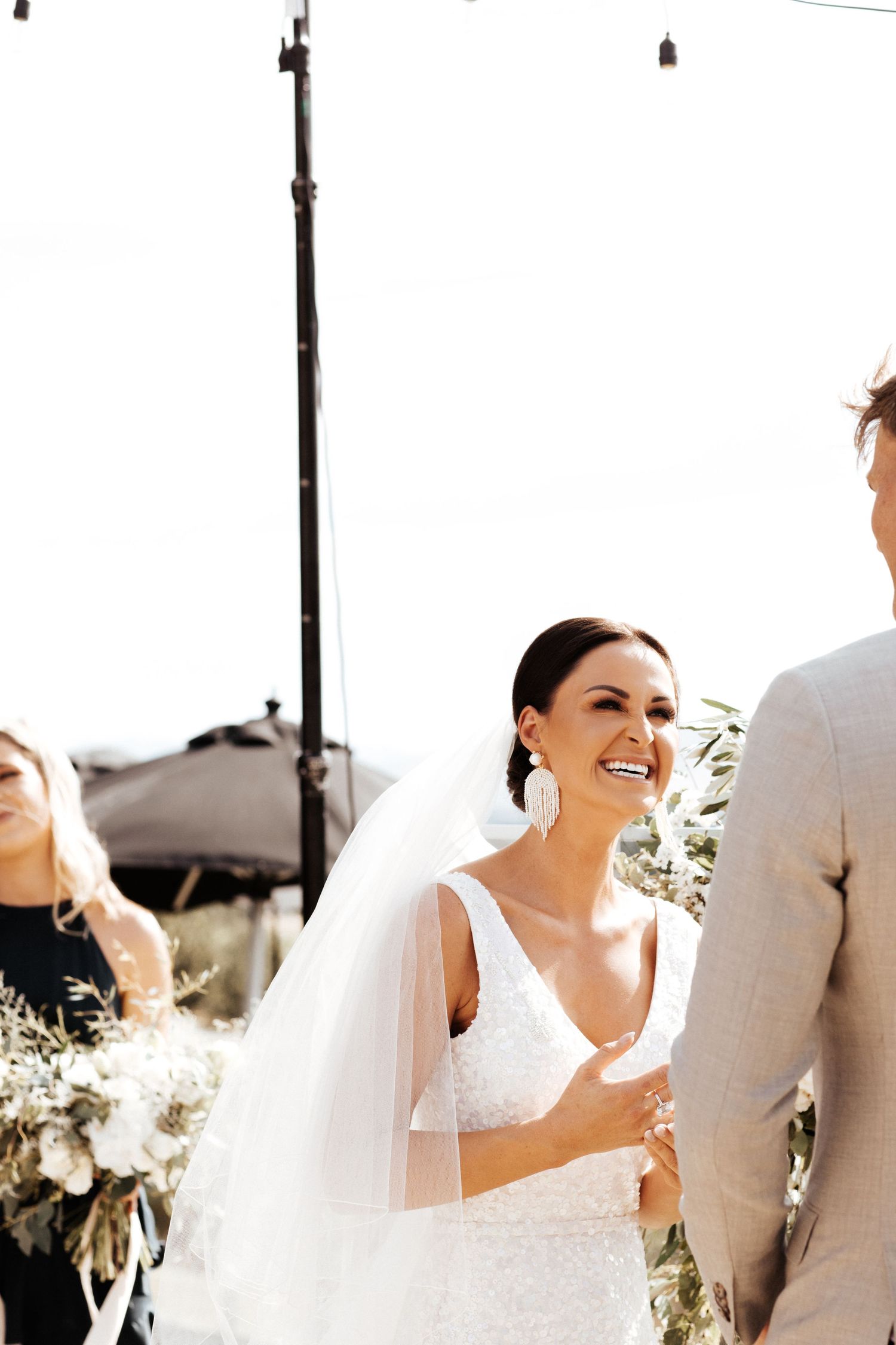 Candid moment of a bride in a white lace dress and veil smiling during her outdoor wedding ceremony.