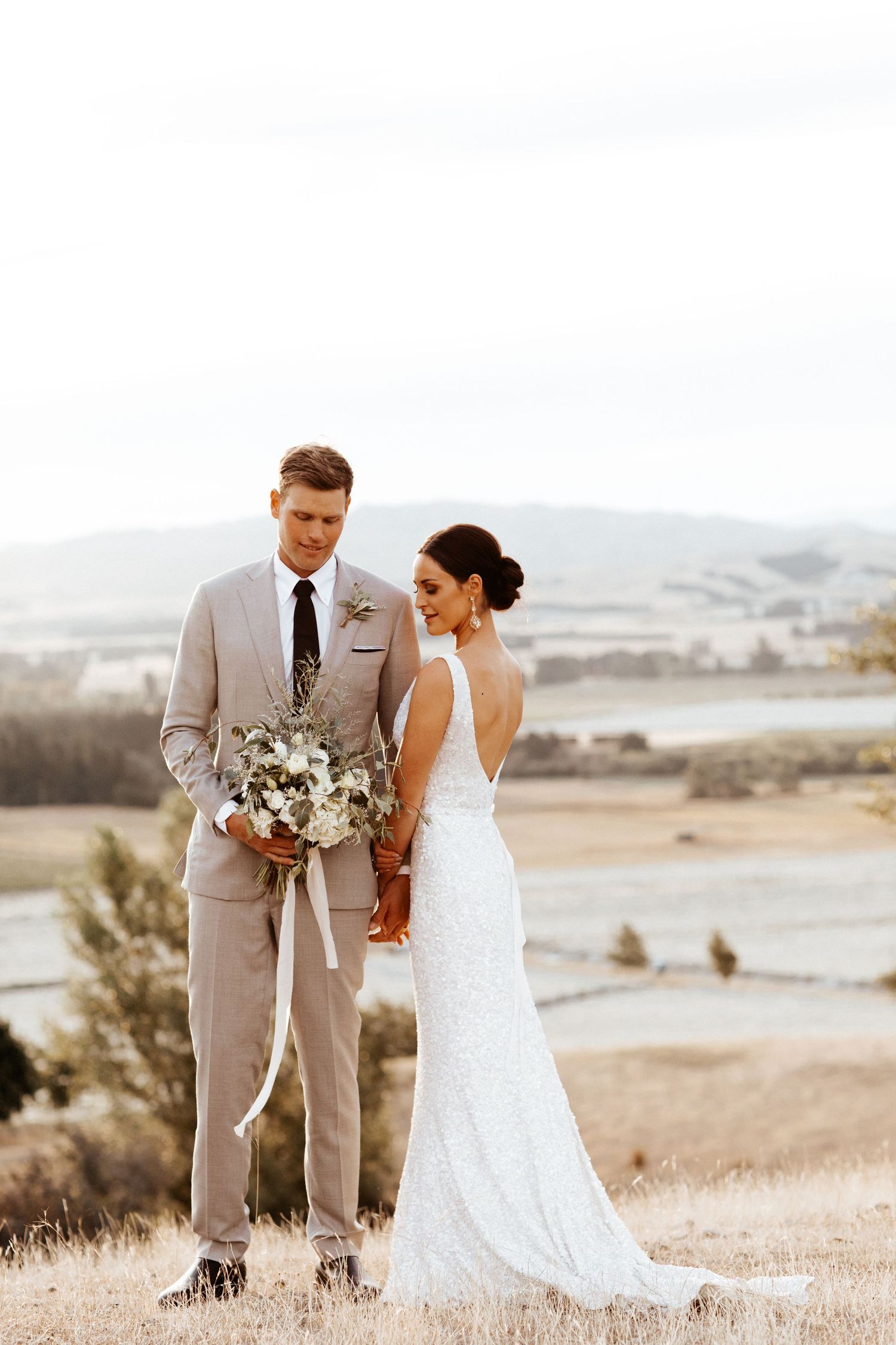Newlyweds stand in a romantic outdoor hilltop setting wearing a light beige suit and white lace wedding dress.