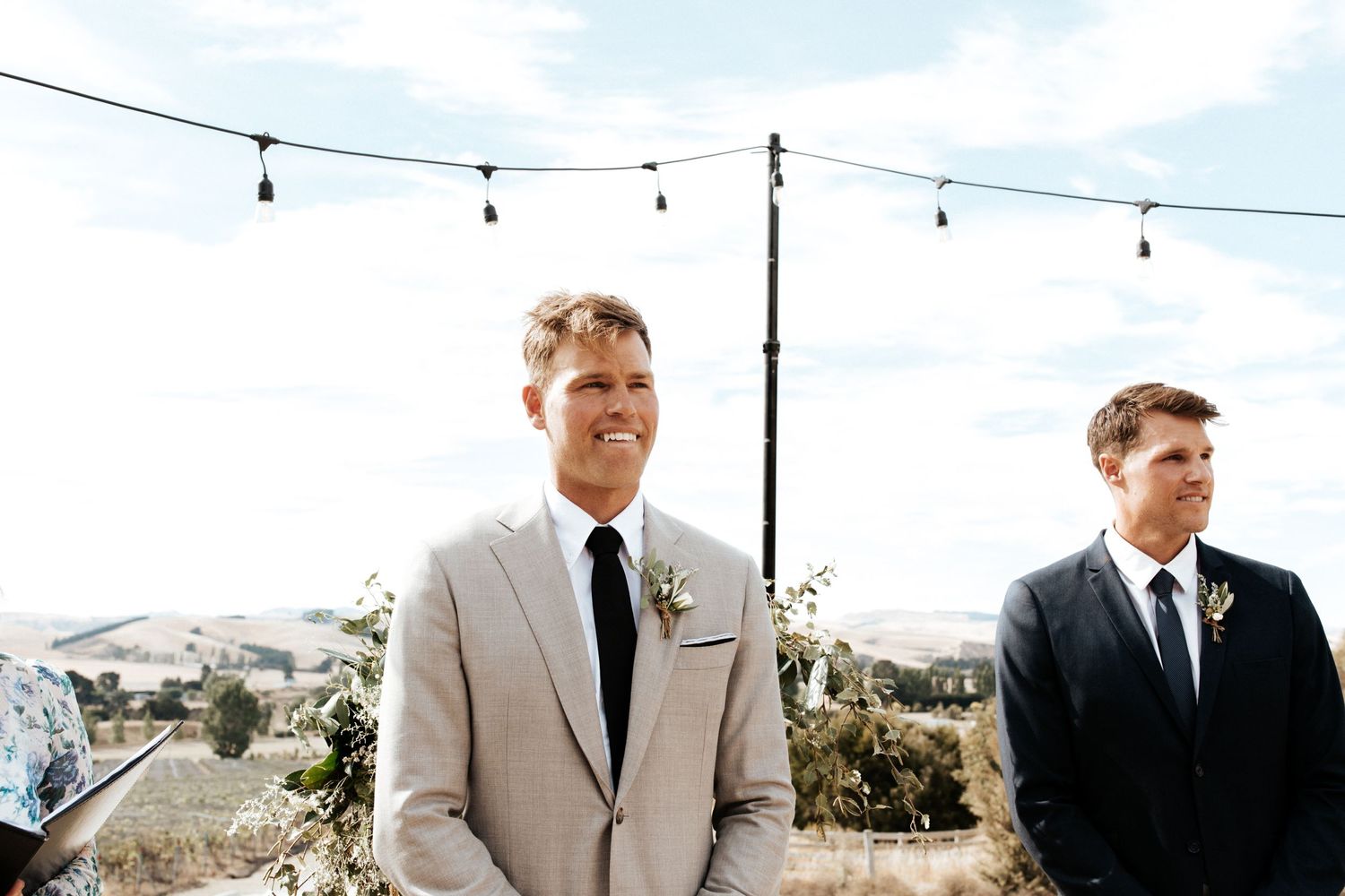 Two groomsmen standing under string lights with mountain landscape behind them.