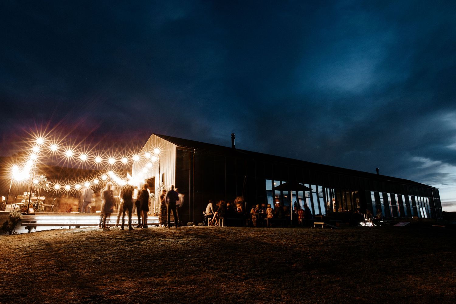 Silhouettes of people gather outside a barn-style building illuminated by warm string lights at night.