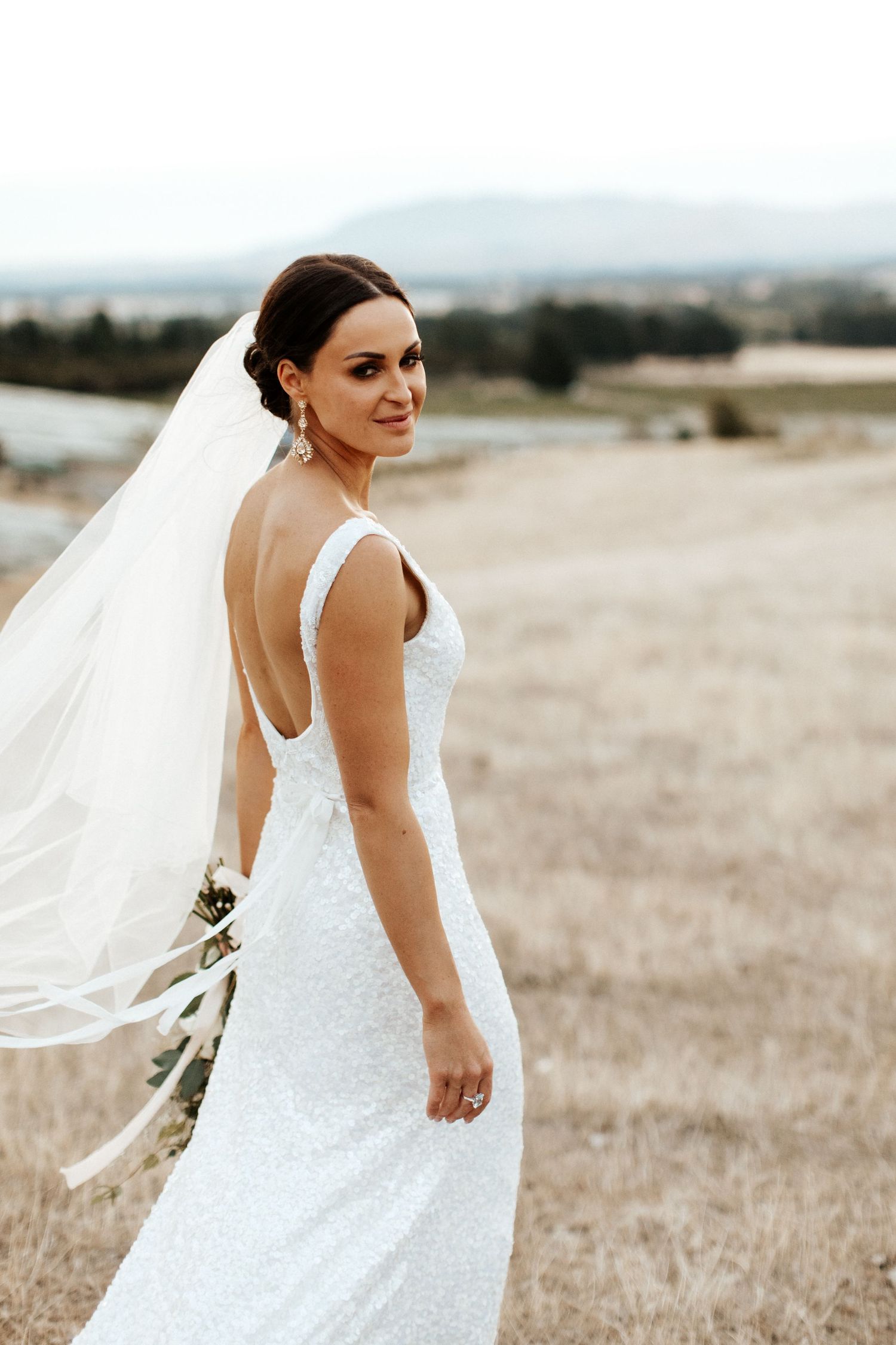 Elegant bride in fitted white dress with low back stands in golden field with flowing veil.