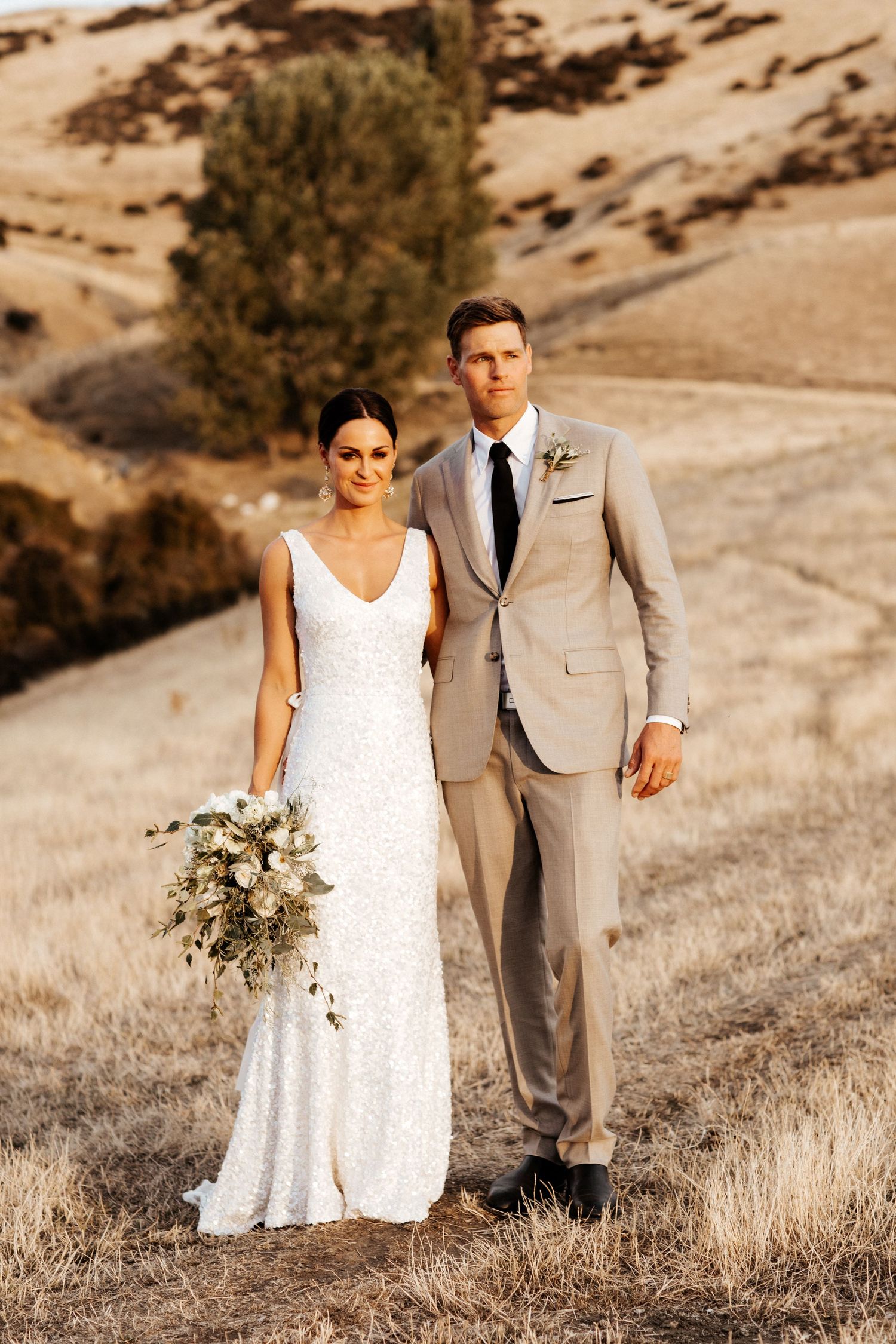 Romantic wedding portrait at golden hour with couple in classic attire standing on rolling hills.