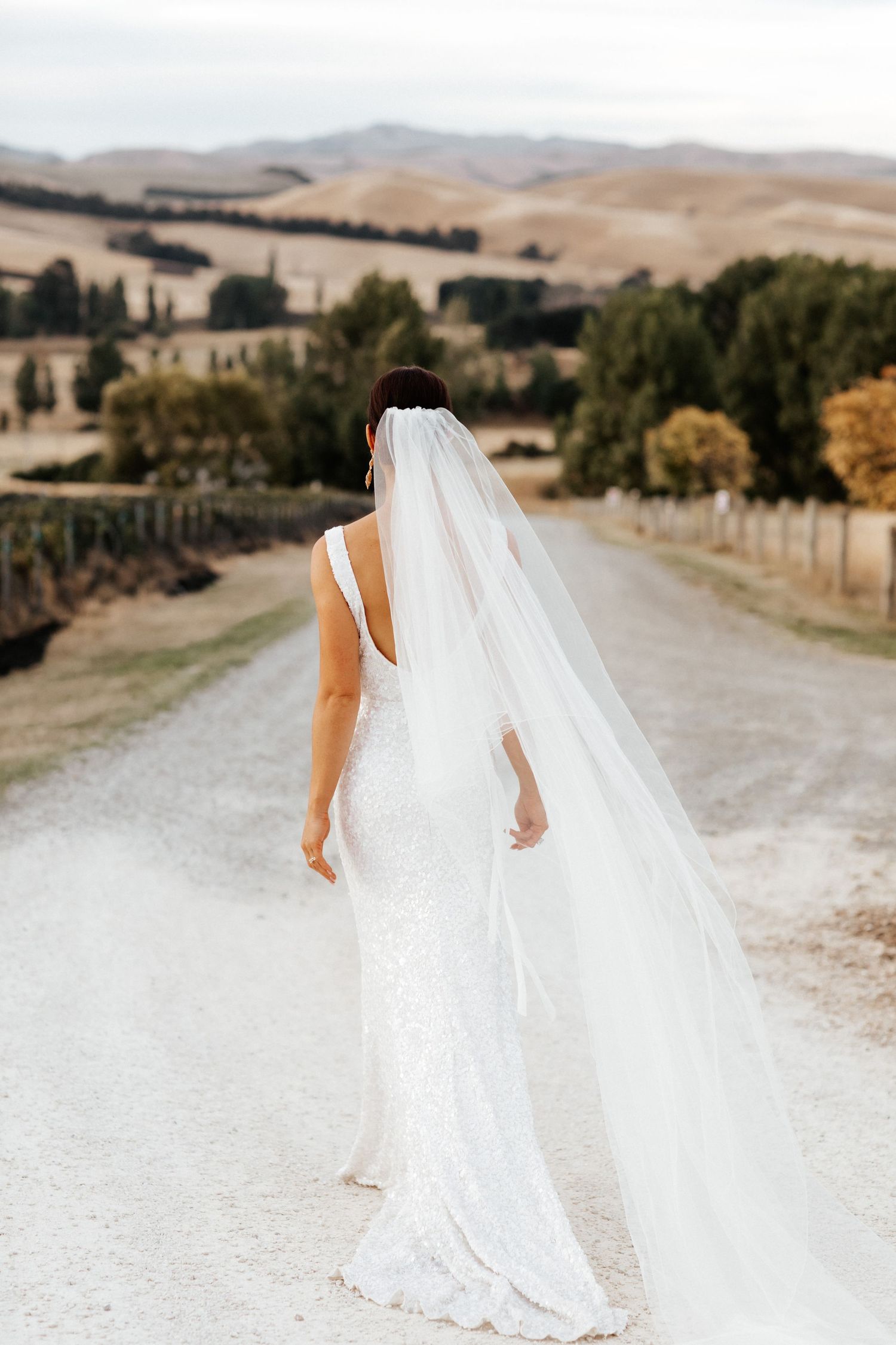 Bride in flowing white dress and long veil walks along countryside path with rolling hills.