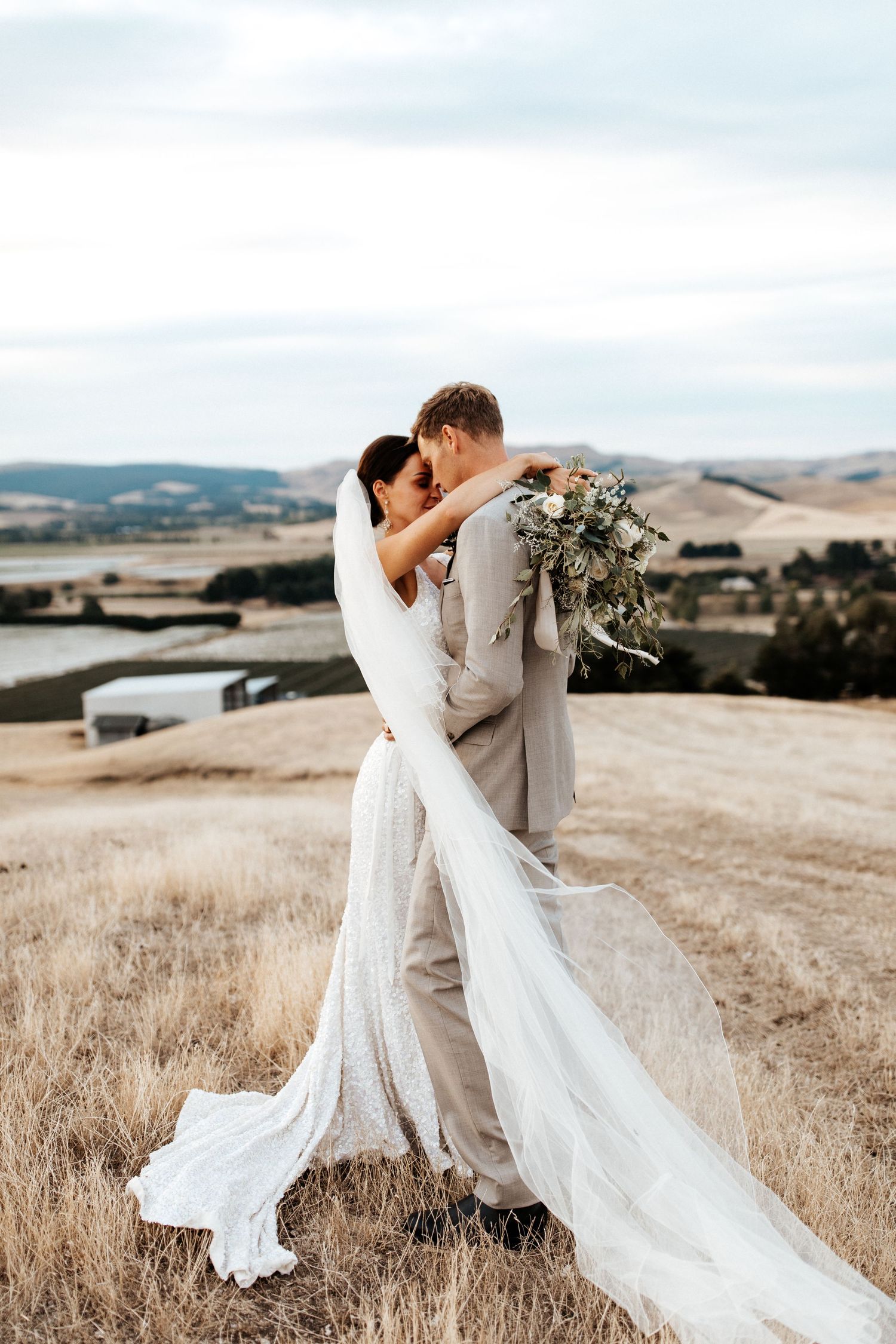 Romantic embrace on hilltop with flowing wedding dress and veil catching the wind.